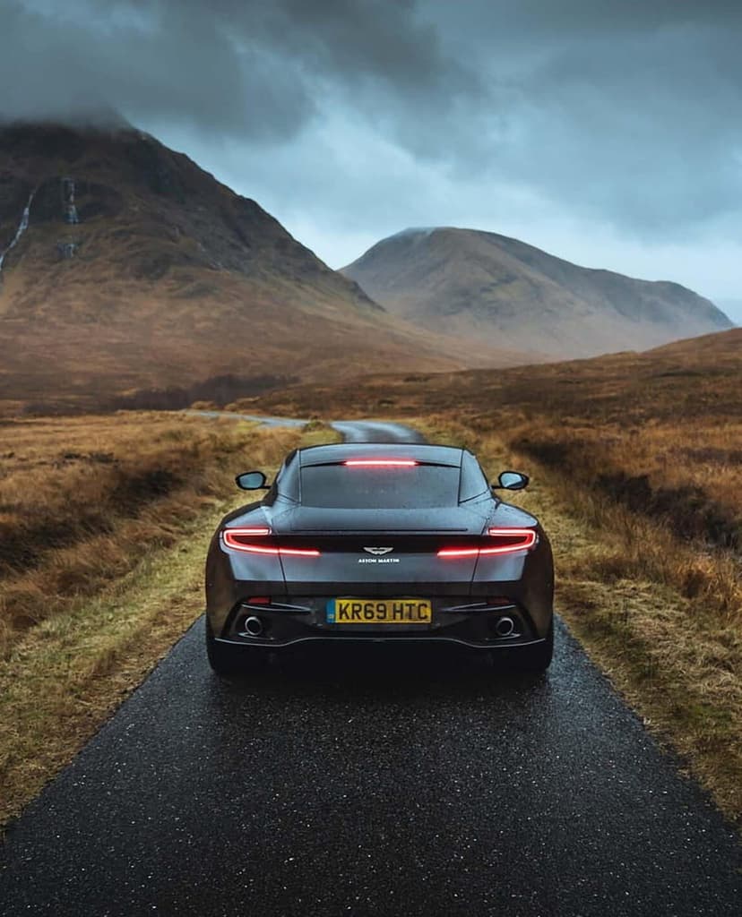 Black Aston Martin with glowing red taillights drives on a wet winding road through misty Scottish mountains.