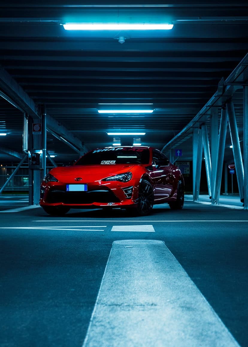 Red sports car parked in a dark concrete garage illuminated by glowing blue overhead fluorescent light strips.