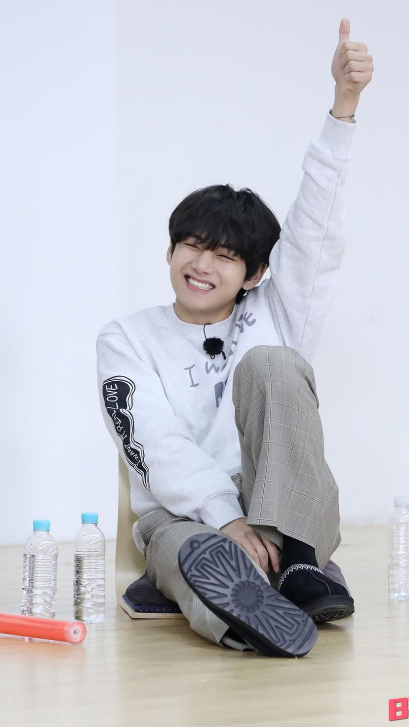 Kim Taehyung smiles and gives a thumbs up while sitting on the floor wearing a white sweatshirt and plaid pants.