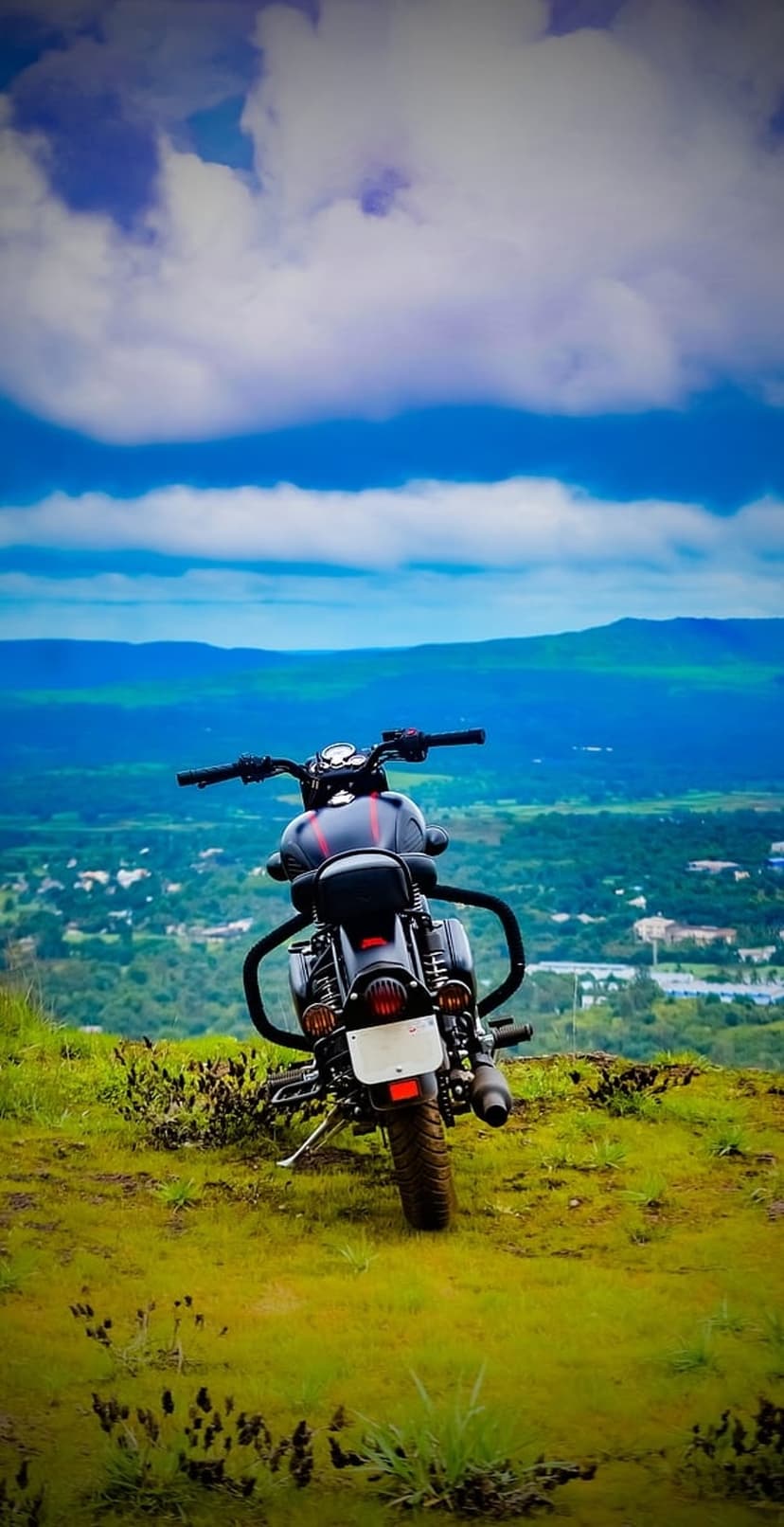 Black Royal Enfield motorcycle rear view on a grassy hilltop overlooking a vast landscape under a blue sky.