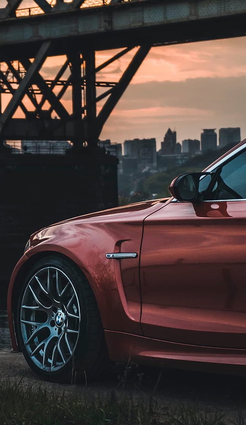 Red BMW fender and wheel reflect sunset light under a metal bridge with a city skyline silhouette in the distance.