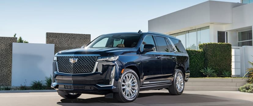 Dark blue Cadillac Escalade SUV parked in front of a minimalist white villa with large windows and a clear sky.