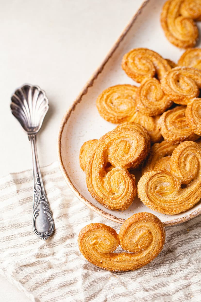 Golden palmier cookies with sugar-coated swirls sit on a white plate next to a vintage silver spoon and napkin.