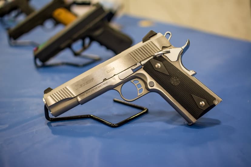 Polished silver 1911 handgun with black grips rests on a display stand against a blurred blue background.