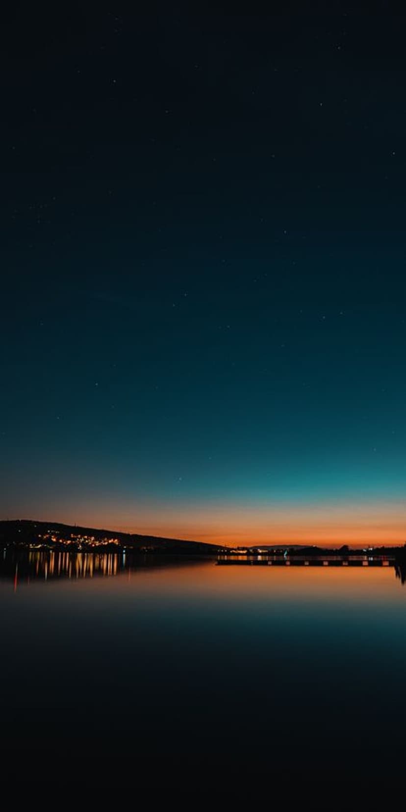 Still lake water reflects a vibrant orange horizon and deep blue sky under a few faint stars at nightfall.
