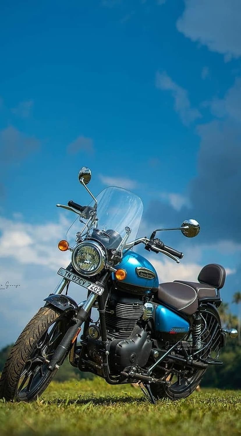 Blue Royal Enfield motorcycle with a chrome engine parked on a lush green lawn under a sunny sky with clouds.
