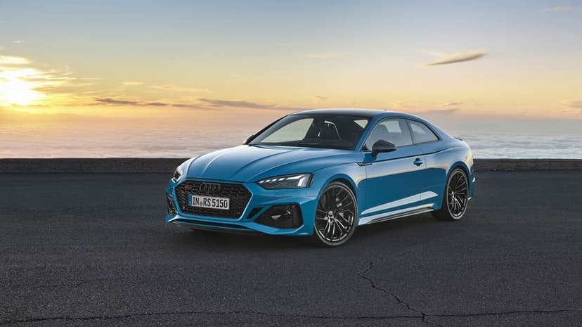 Blue Audi RS5 Coupe parked on a high asphalt road at sunset overlooking a vast sea of clouds with golden lighting.