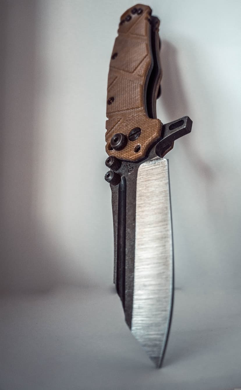 Tactical folding knife with a tan textured handle and sharp satin cleaver blade over a neutral gray backdrop.