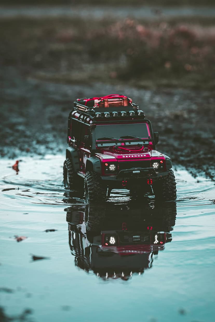 Red scale model SUV with roof rack and off-road tires reflects in a shallow water puddle under moody lighting.