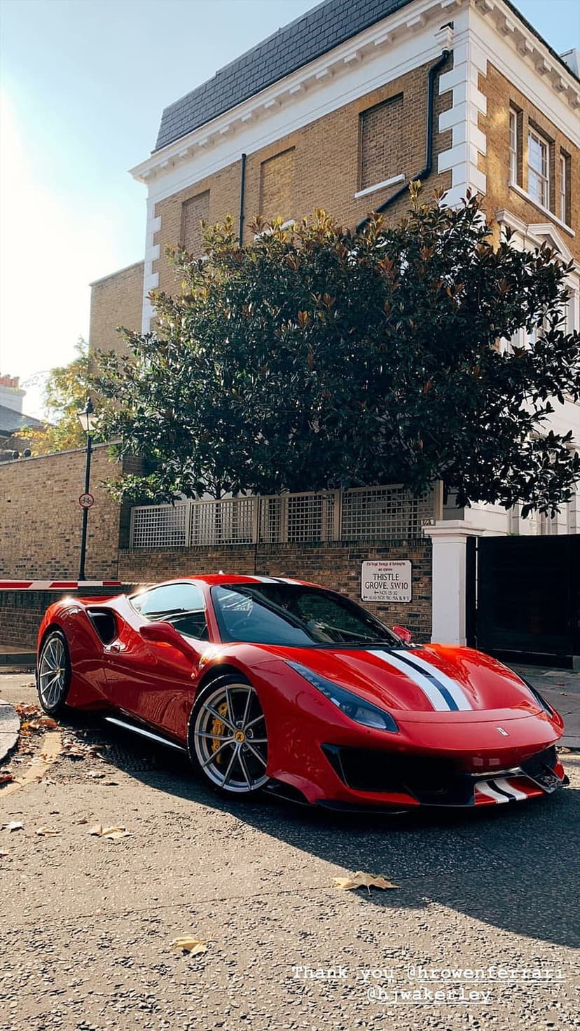 Red Ferrari with racing stripes parked on a London street in front of a classic brick building in sunlight.