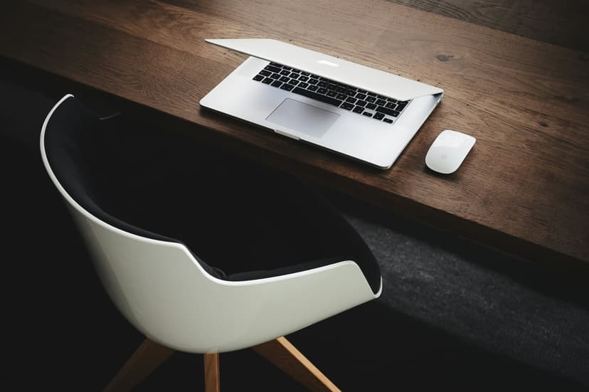 Silver laptop and white wireless mouse rest on a polished wooden desk beside a black and white ergonomic chair.