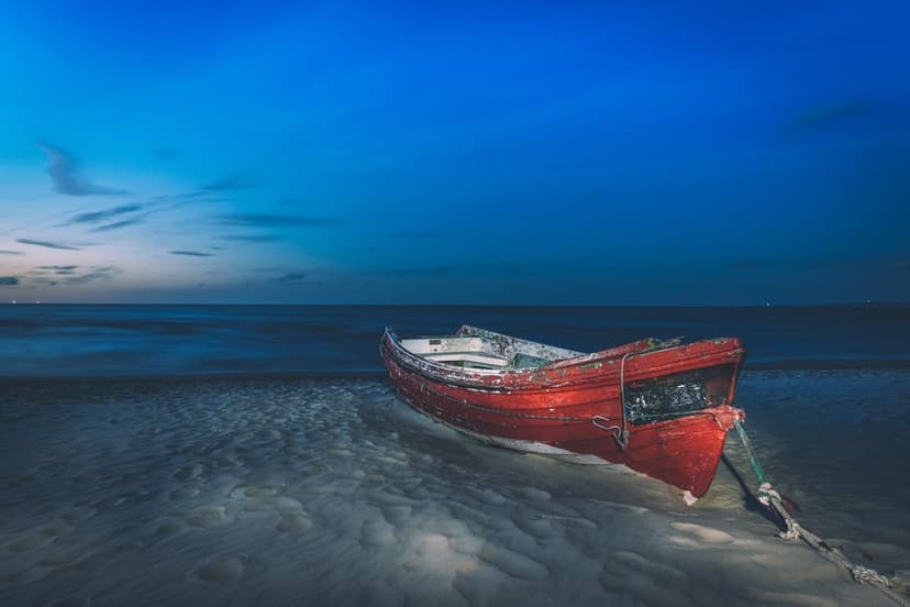 Red wooden boat rests on a sandy shore under a vast navy blue twilight sky with a calm ocean in the distance.