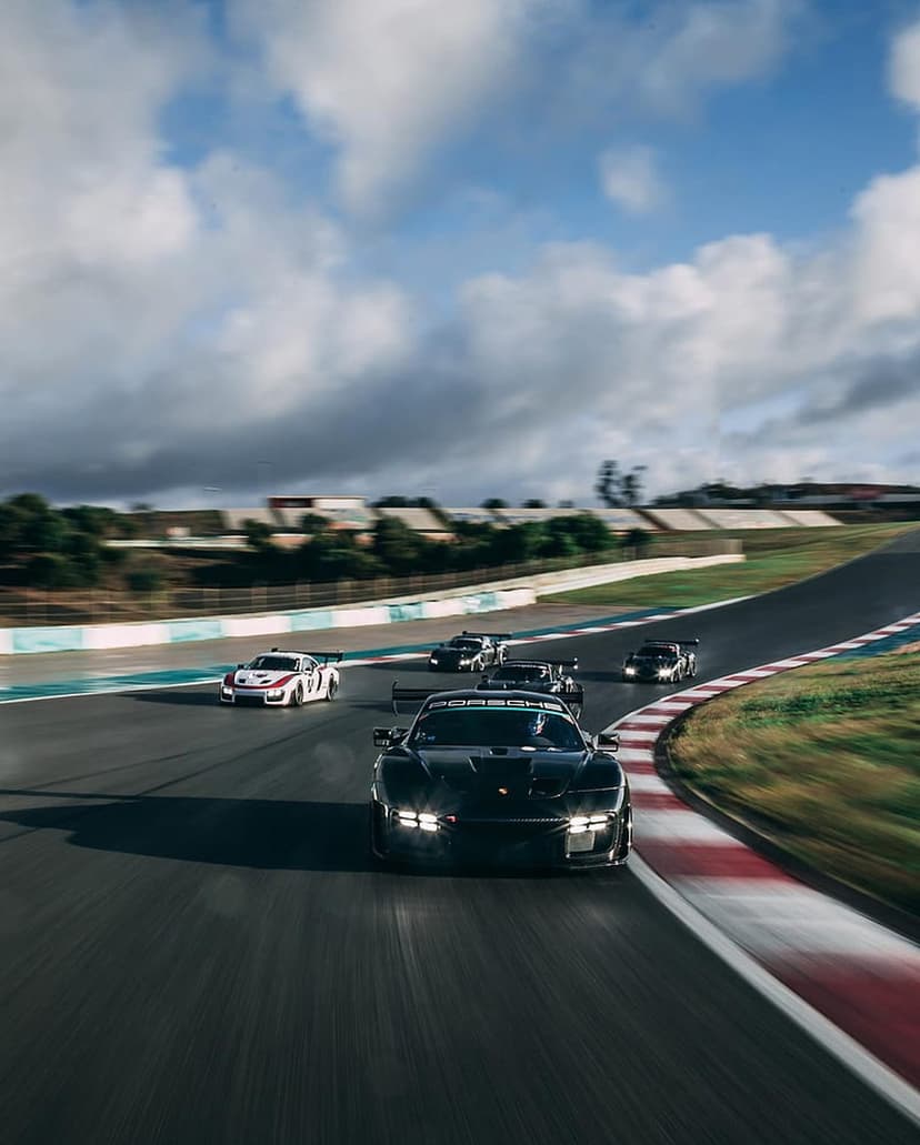 Black Porsche race car with glowing headlights leads a pack of vehicles on a curved racetrack during a cloudy day.