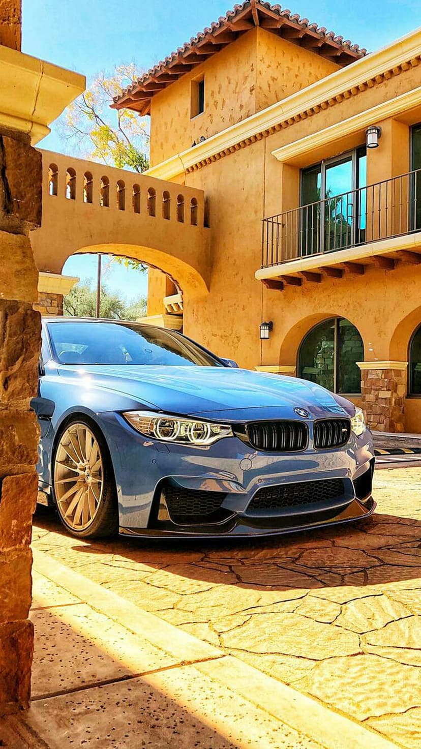 Blue BMW M4 sports car parked on stone pavement in front of a Mediterranean building with arched walkways.