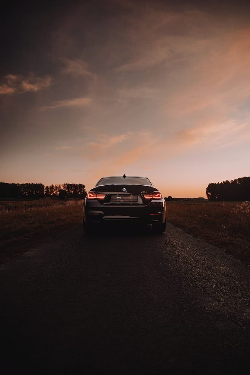 BMW M4 sports car parked on wet asphalt at sunset with glowing taillights and a colorful cloud-filled sky.