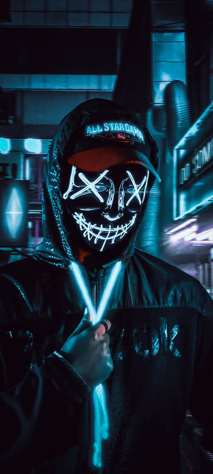 Hooded figure with a glowing X-eyed mask holds a luminous blue orb amidst a neon-lit futuristic cityscape.