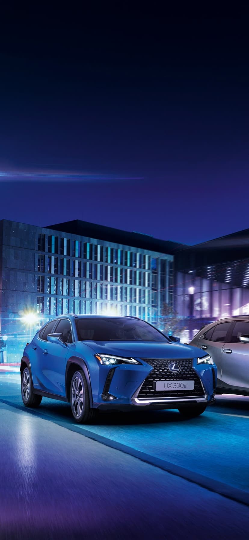 Blue Lexus UX 300e electric SUV driving on a city street at night with motion blur and architectural lights.