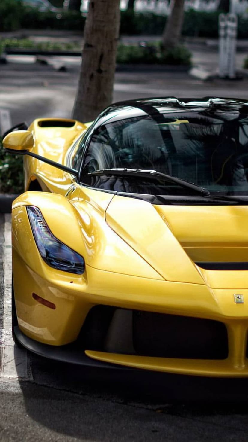 Yellow Ferrari LaFerrari parked on asphalt showing its aggressive front end, sleek headlights, and tree reflections.