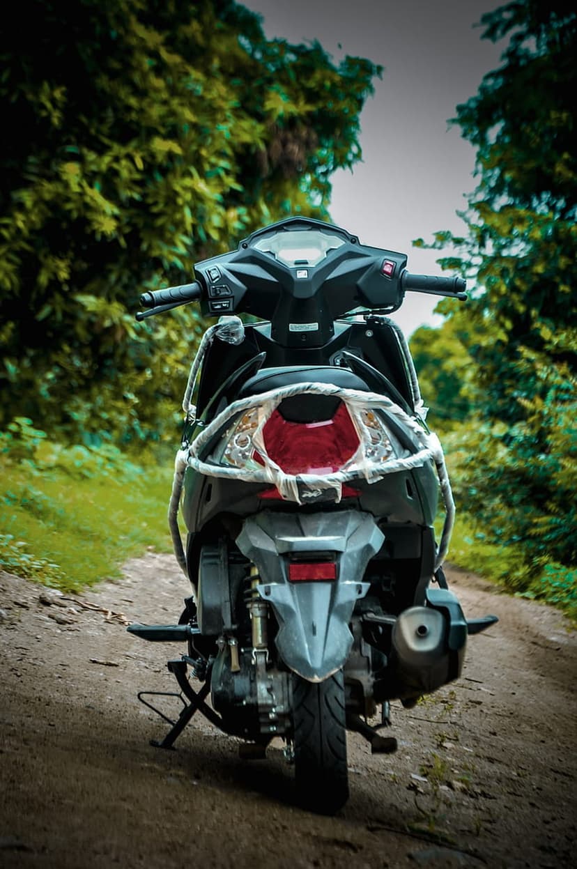 Black scooter with a red taillight parked on a dirt forest path surrounded by dense green trees and foliage.