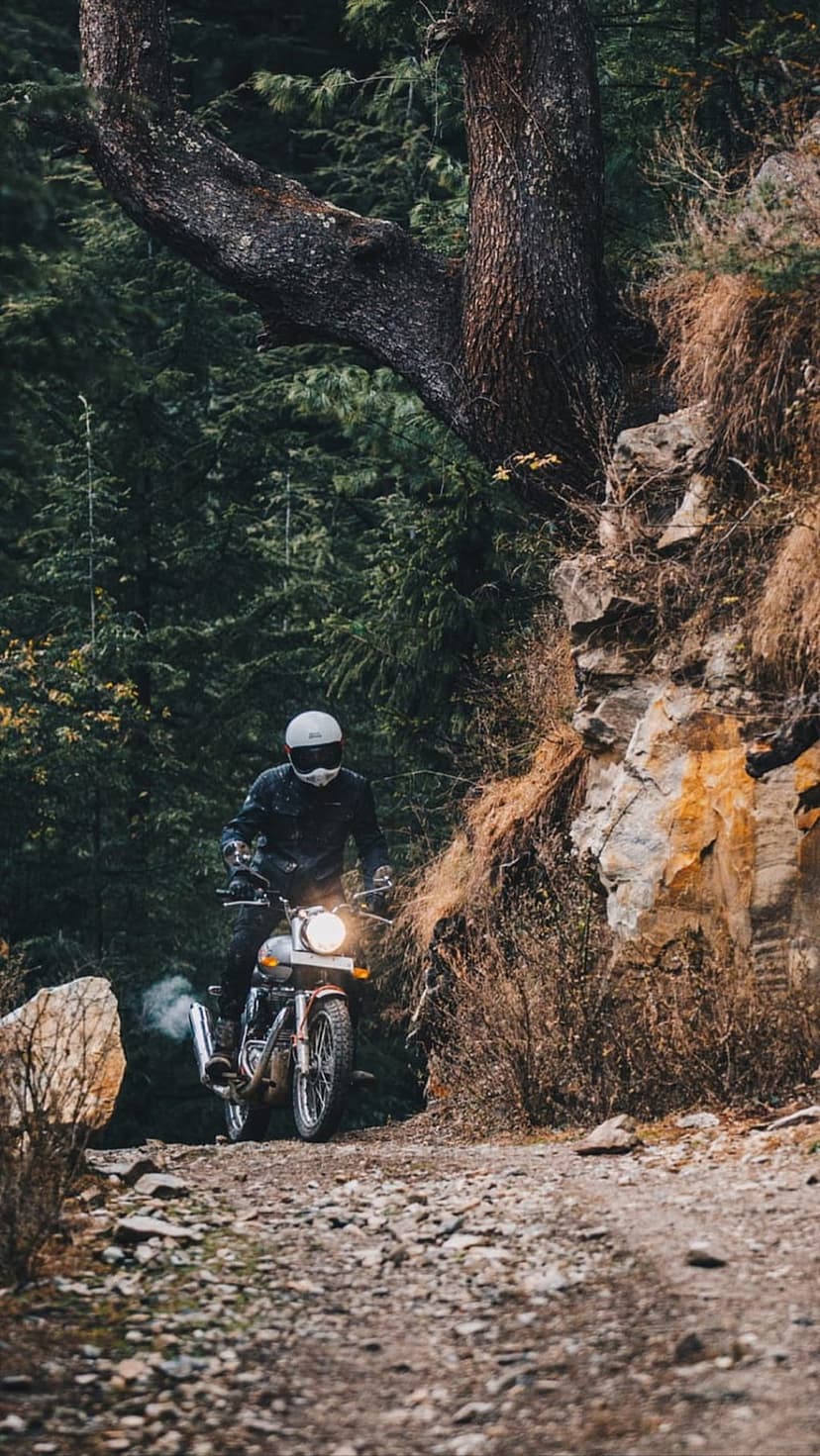 Motorcyclist in a helmet rides a vintage bike on a rocky trail through a sunlit, dense green forest.