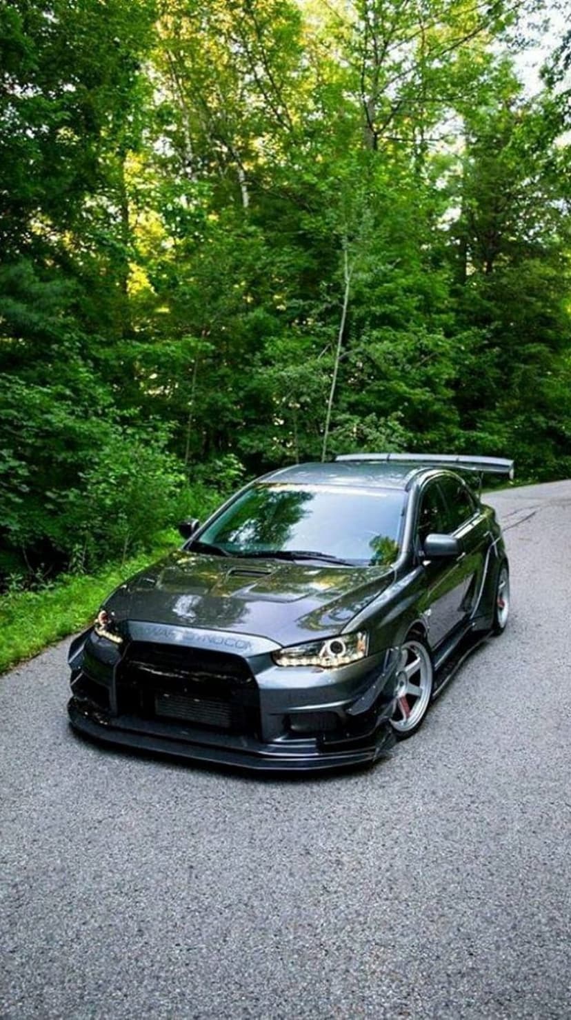 Dark grey Mitsubishi Evo X with a widebody kit and large wing parked on a forest road with glowing headlights.