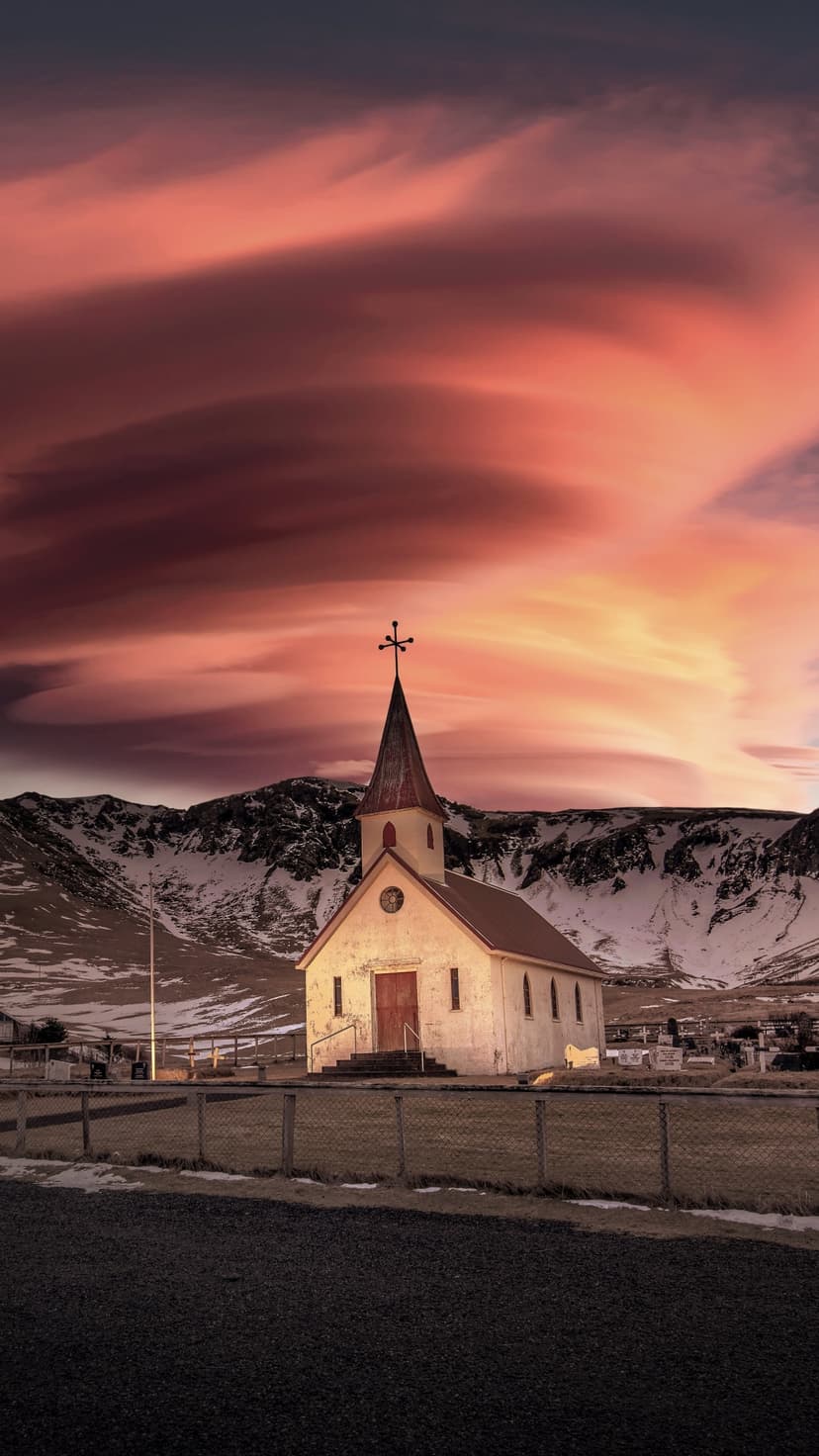 White countryside church with a dark steeple stands before snow-dusted mountains under a pastel sunset.