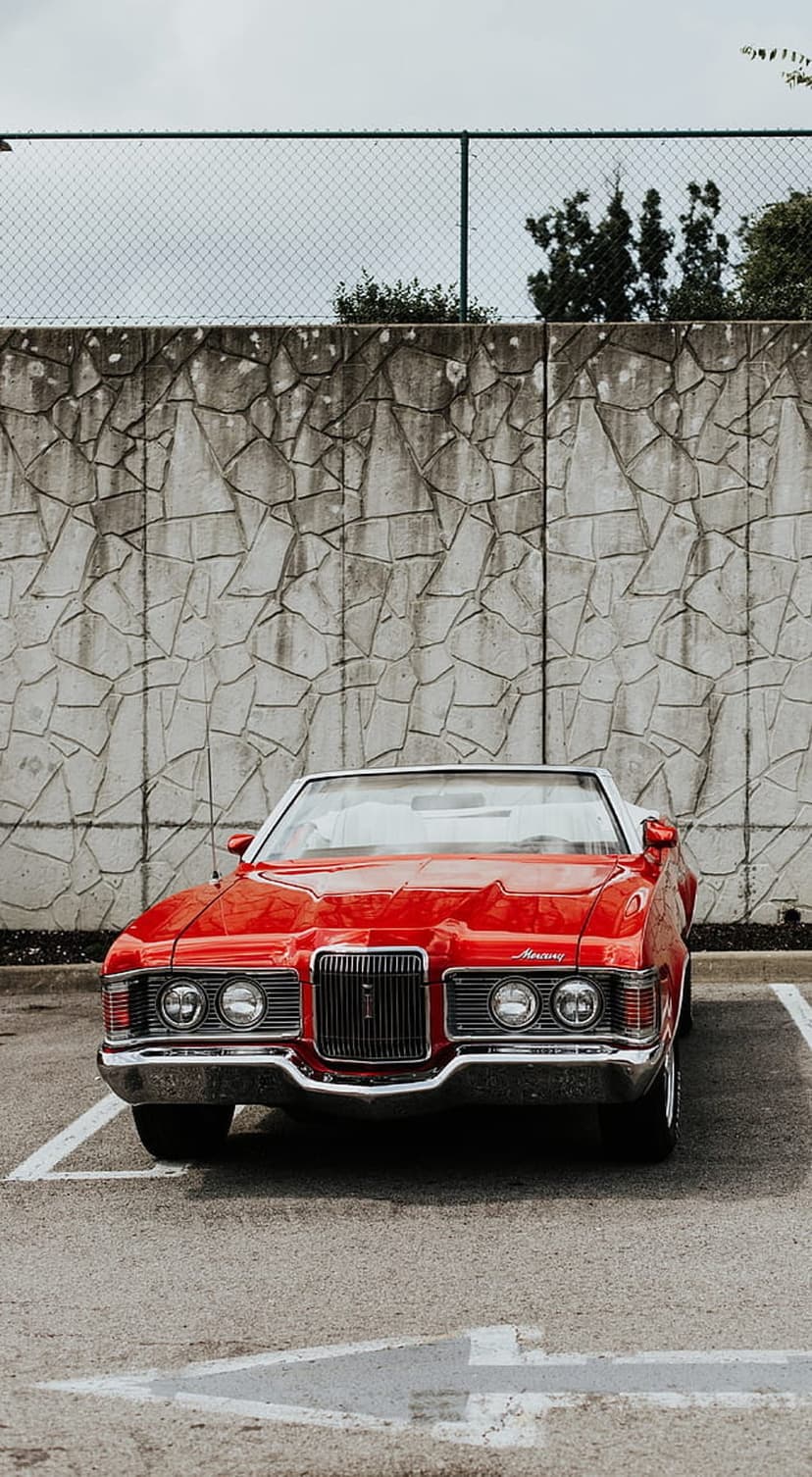 Red vintage convertible car parked in front of a textured stone wall with chrome details and an open top.