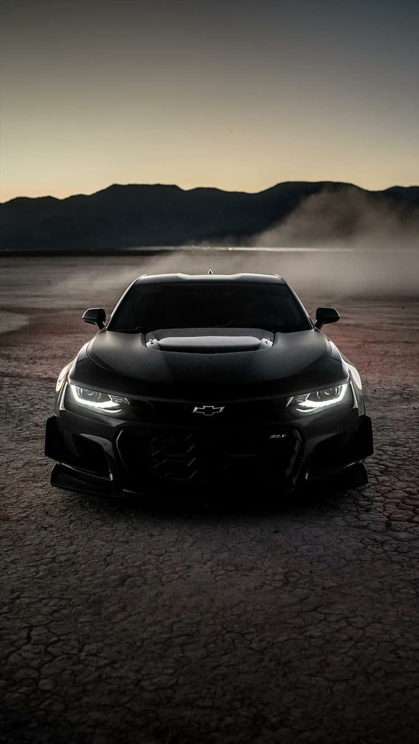 Black Chevrolet Camaro ZL1 with glowing headlights on cracked desert soil under a hazy mountain twilight sky.