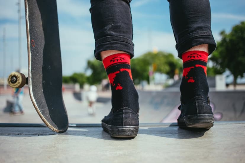 Skateboarder's legs in red patterned socks and black sneakers at a sunny skatepark with a blurred background.