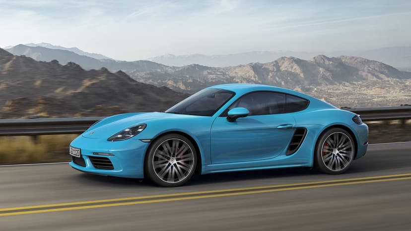 Light blue Porsche 718 Cayman driving fast on a desert road with rugged mountains in the background.