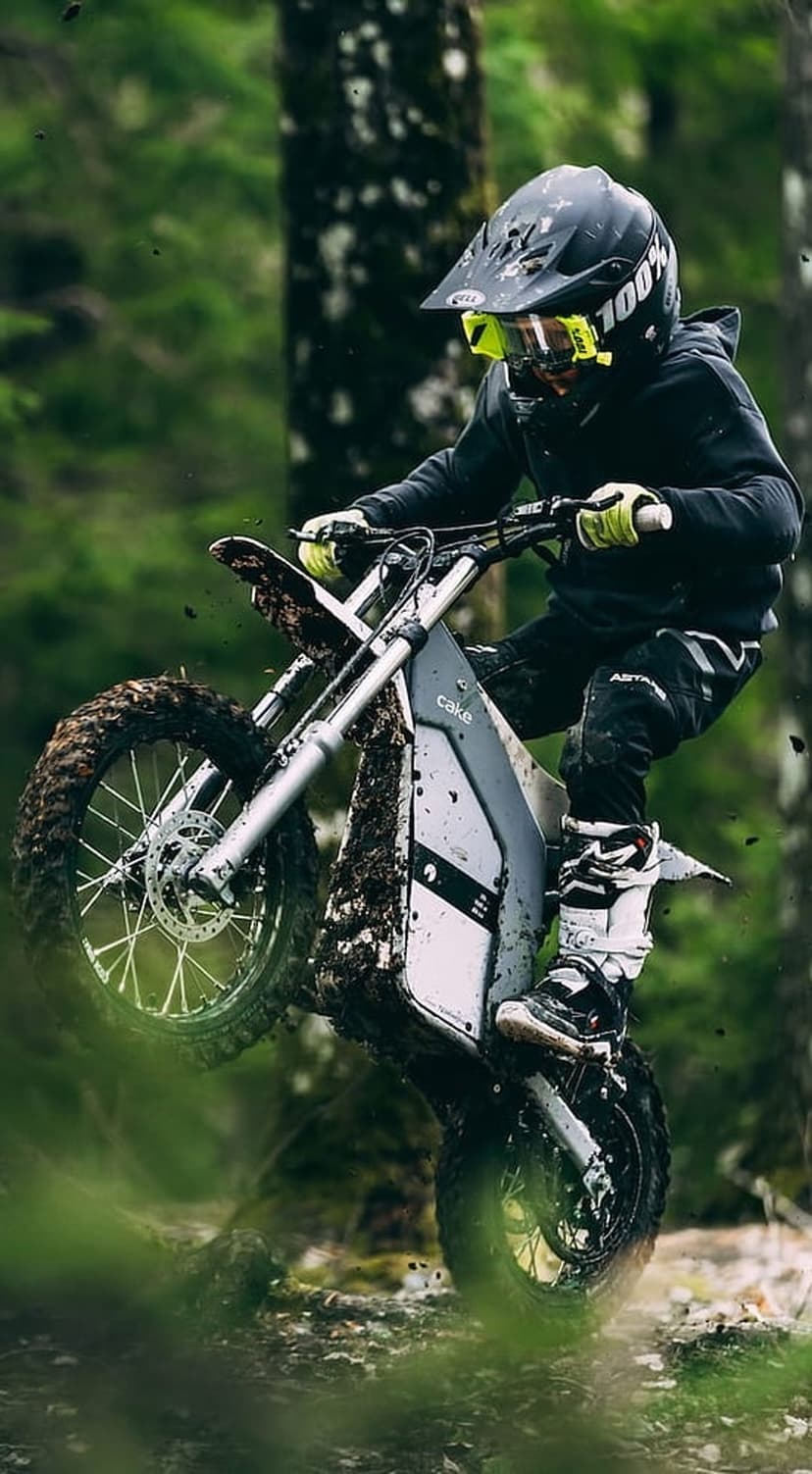Motocross rider on a Cake electric bike kicks up mud while navigating a wet, forested off-road trail in motion.
