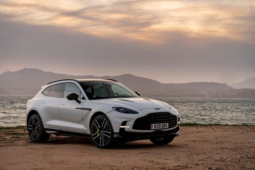 White Aston Martin DBX parked on a sandy beach at sunset with mountains and water in the background.