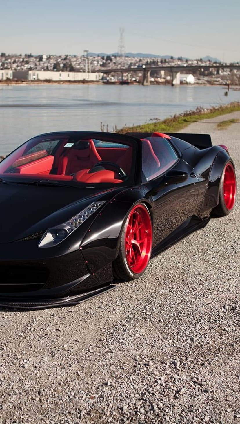 Black Ferrari convertible with red interior and rims parked by water, featuring a bridge and city skyline.