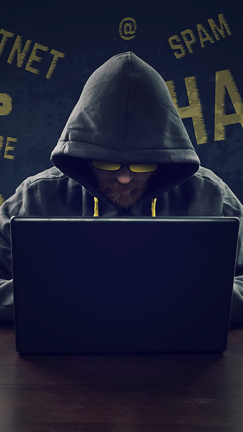 Person in a dark hoodie and yellow glasses hunched over a glowing laptop screen in a low-light room.