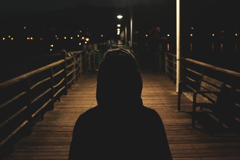 Hooded figure silhouetted on a wooden pier against a distant skyline of glowing city lights and dark water.