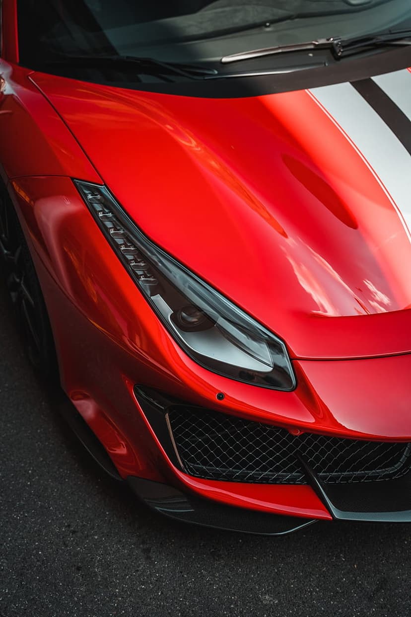 Red Ferrari headlight and racing stripes on a glossy hood, showcasing metallic reflections in close-up detail.