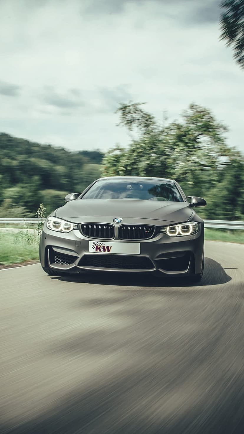 Silver BMW M4 sports car takes a sharp turn on a tree-lined road with visible motion blur and a low-angle view.