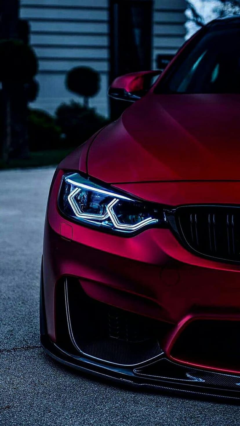 Crimson sports car headlight with glowing LED accents and matte red bodywork reflected against dark asphalt.