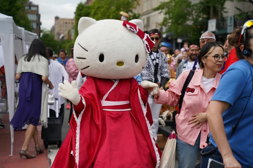Hello Kitty mascot in a red kimono stands before a crowded street festival with white tents and a diverse parade.