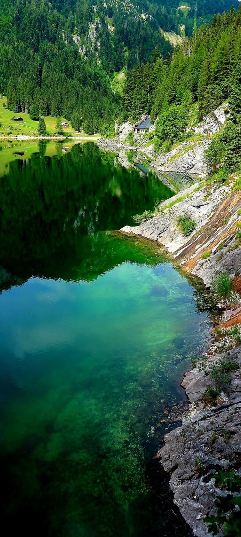 Emerald lake water reflects a green pine forest and a small house on a rocky mountainside in a vertical view.