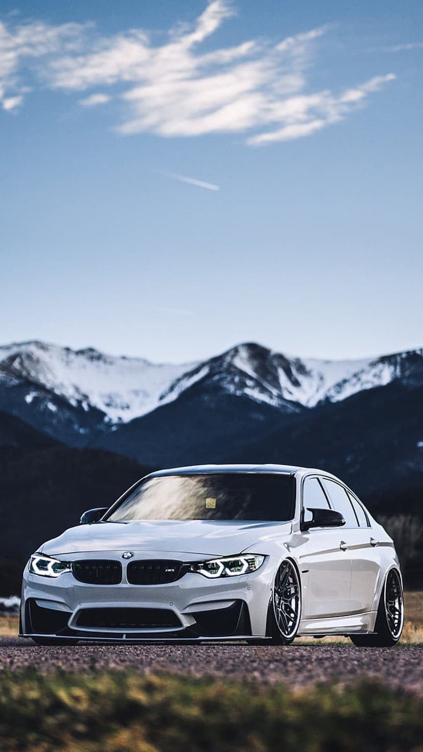 White BMW M3 sedan parked on a mountain road with jagged snow-covered peaks under a clear blue winter sky.