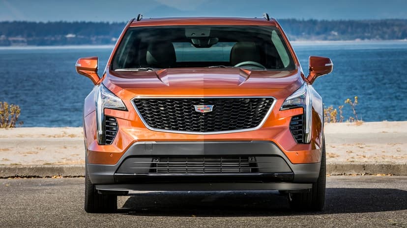 Front view of a metallic orange Cadillac SUV parked on asphalt against a blue ocean and mountain backdrop.