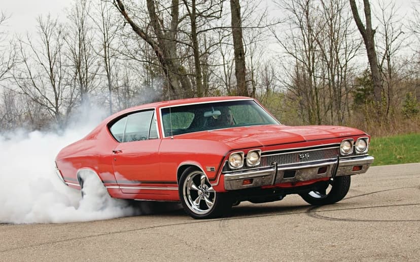 Red classic muscle car performing a smoky burnout on a paved road with thick white tire smoke and chrome accents.