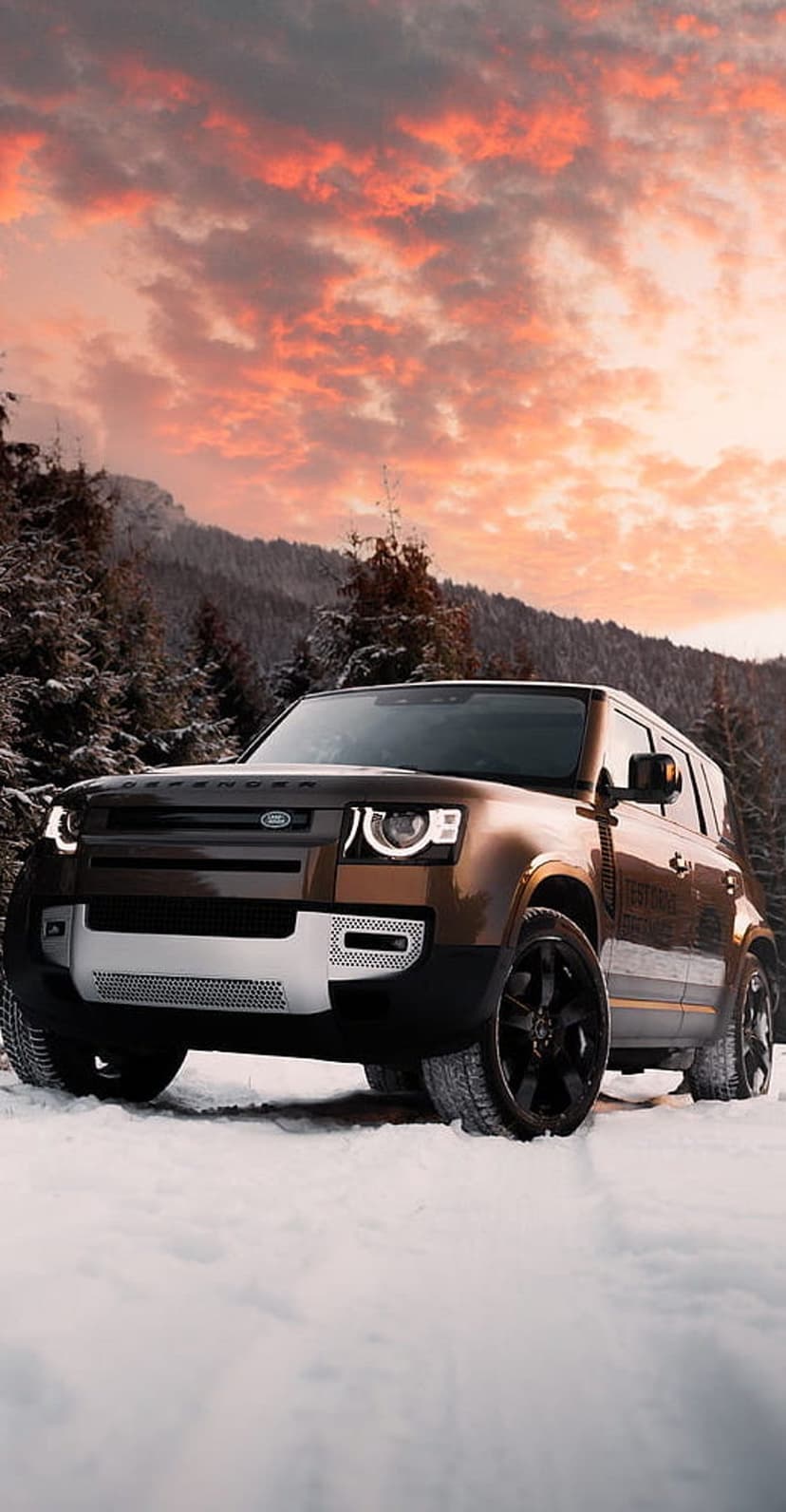 Land Rover Defender SUV parked in deep snow against glowing orange clouds and jagged winter mountains.