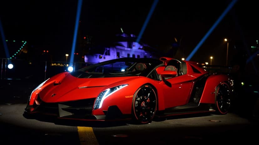 Red Lamborghini Veneno Roadster parked on a dark flight deck with blue spotlights and a helicopter background.