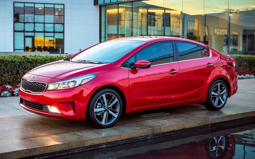 Red Kia Forte sedan parked by a modern glass building with reflections and a calm pool in the foreground.