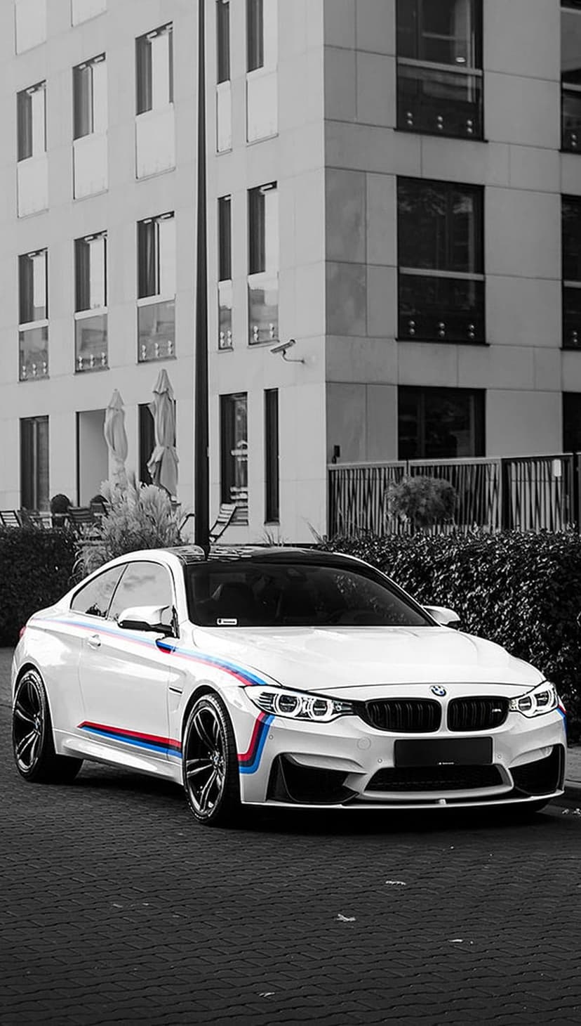 White BMW M4 with red and blue stripes parked on dark pavement facing a monochrome multi-story building.