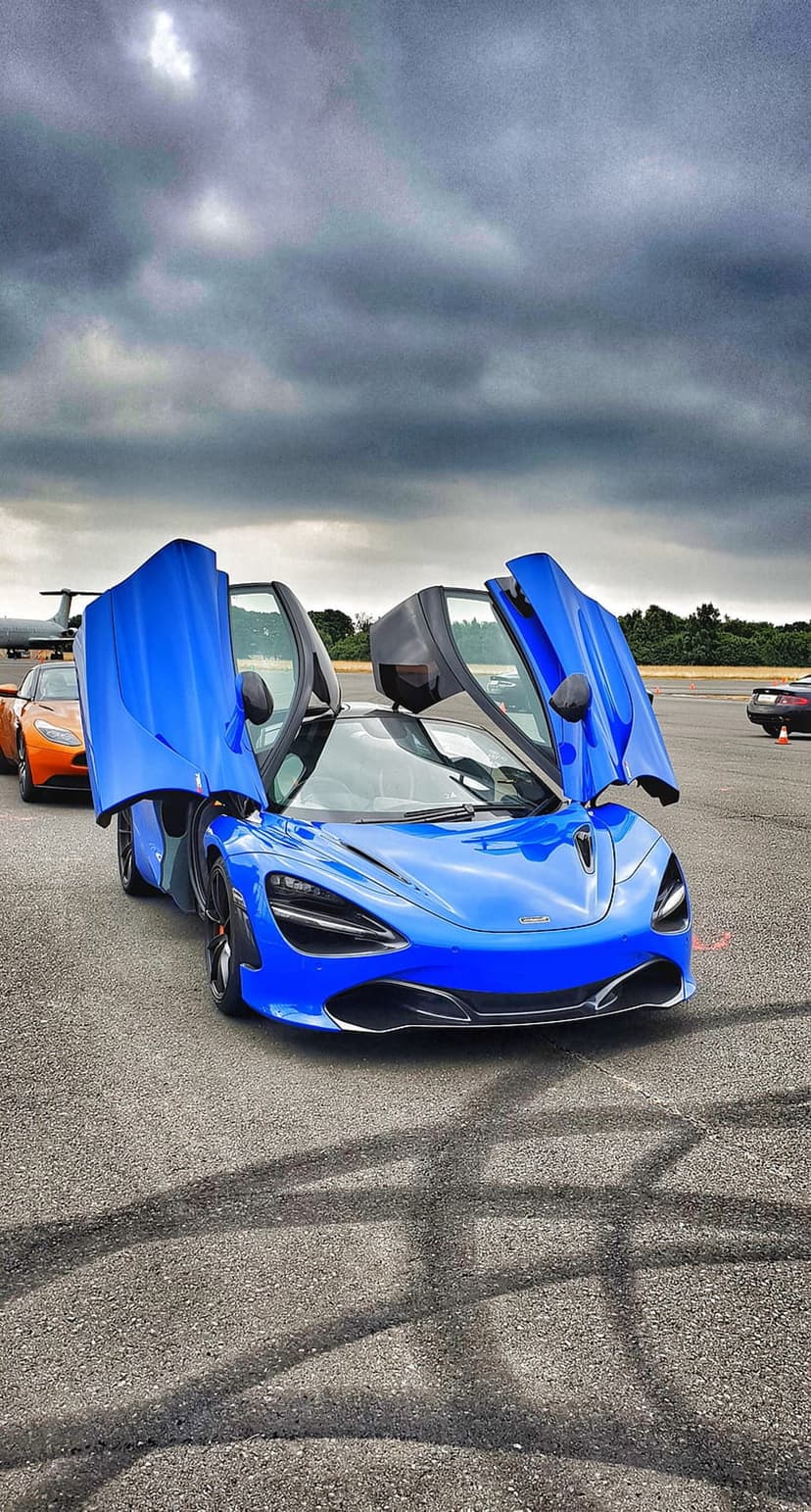 Blue McLaren 720S supercar with butterfly doors open on a textured asphalt racing track under a cloudy sky.