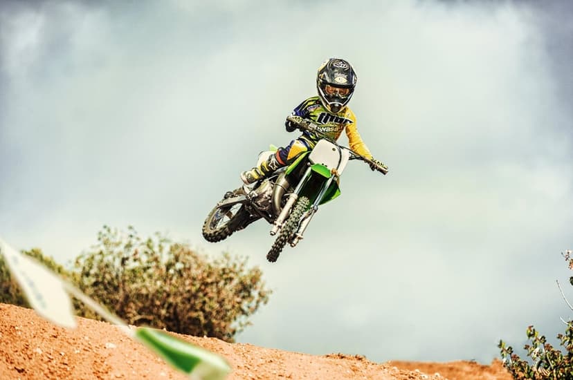 Young motocross rider in yellow gear jumps a green dirt bike high against a cloudy sky above a dirt track.