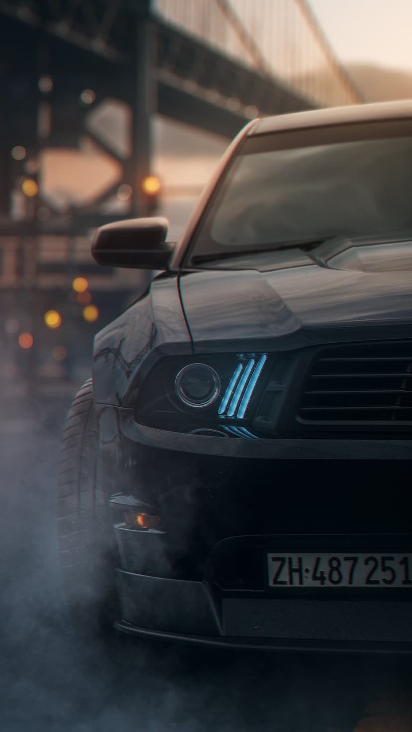 Mustang headlights with blue LED strips glow through street smoke against a bokeh urban background at night.